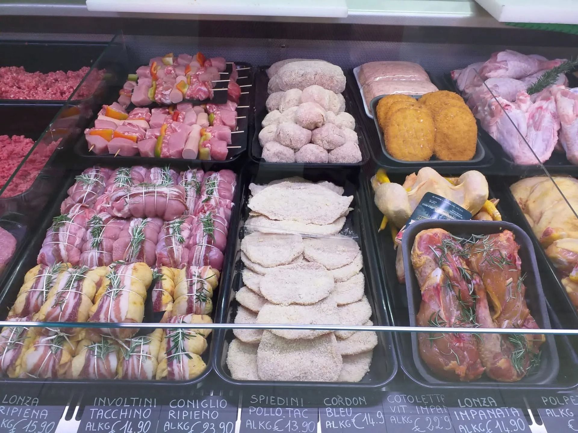 Una vetrina da banco gastronomia piena di vassoi con vari tagli di carne cruda, spiedini, prodotti impanati e pollame condito.