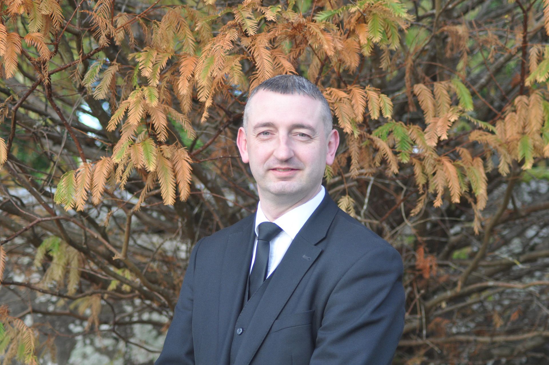 Peter Lowden in a suit and tie is standing in front of a tree.