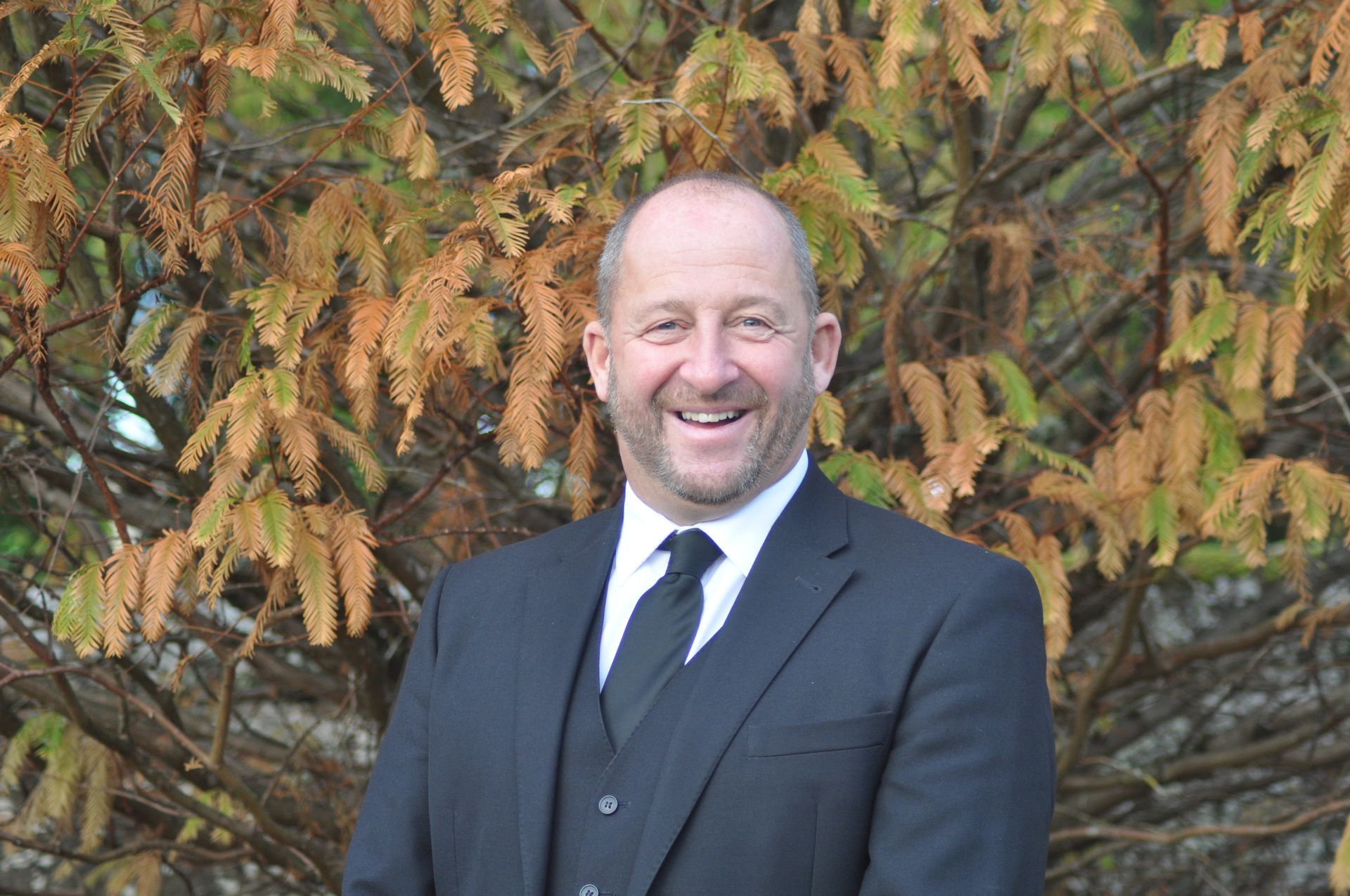 John Stewart in a suit and tie is smiling in front of a tree.