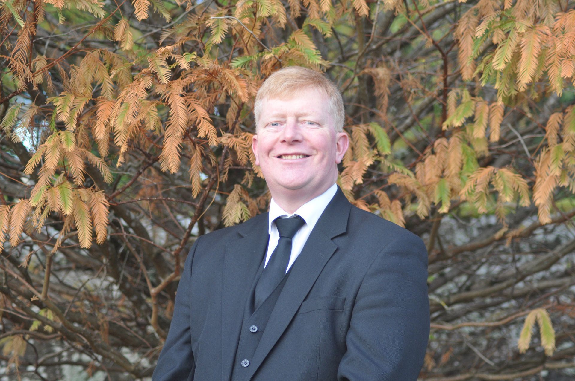 Bruce Dow in a suit and tie is standing in front of a tree.