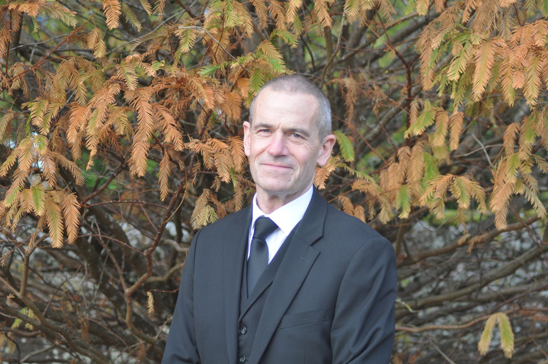 John Stuart in a suit and tie is standing in front of a tree.