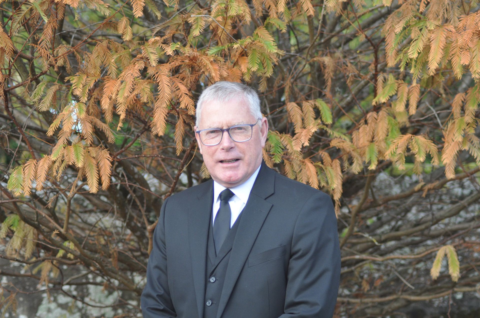 Billy Mackay in a suit and tie is standing in front of a tree.
