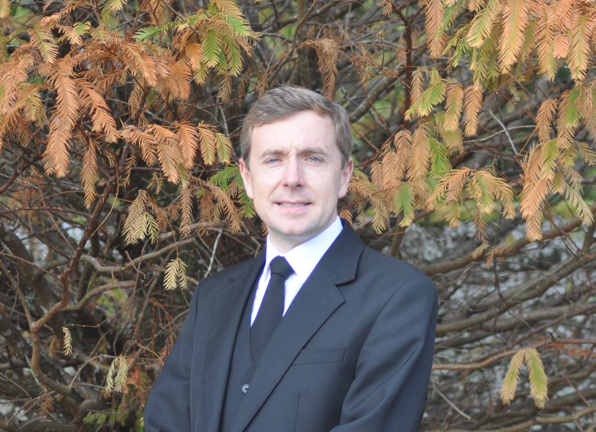 Martin Mackay in a suit and tie is standing in front of a tree.