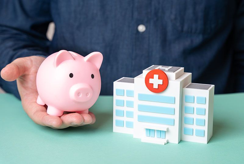 A hand holds a pink piggy bank next to a small, white model of a hospital building on a light green surface.