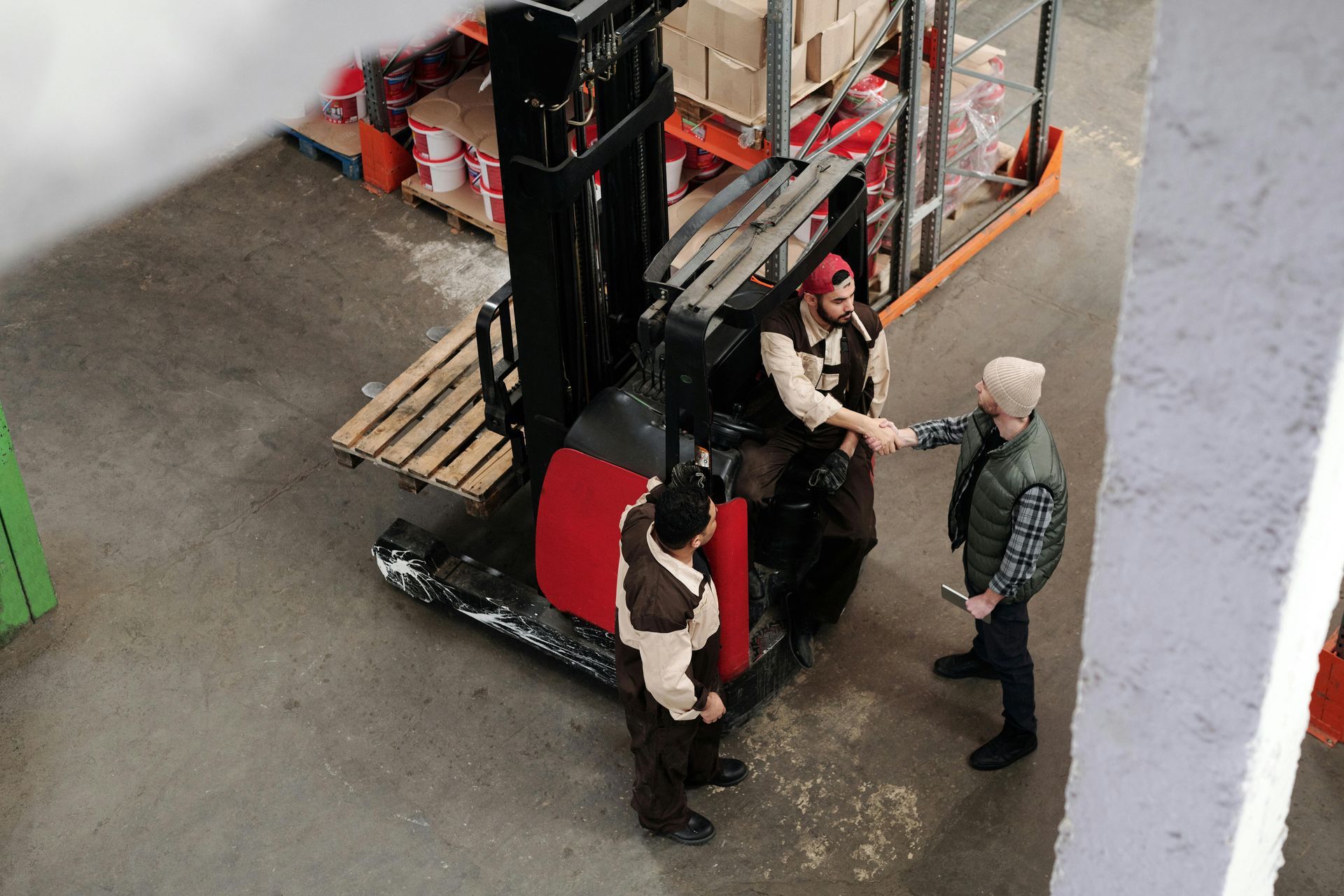 Three people stand in a warehouse, with two shaking hands near a parked forklift while a third person watches.