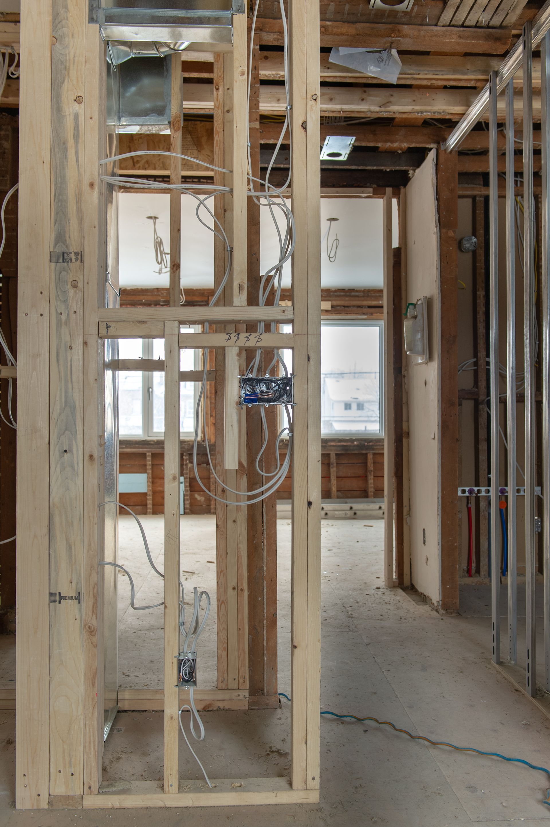 Interior view of a building under construction, showing exposed wooden framing, electrical wiring, and a doorway leading to a room with windows.