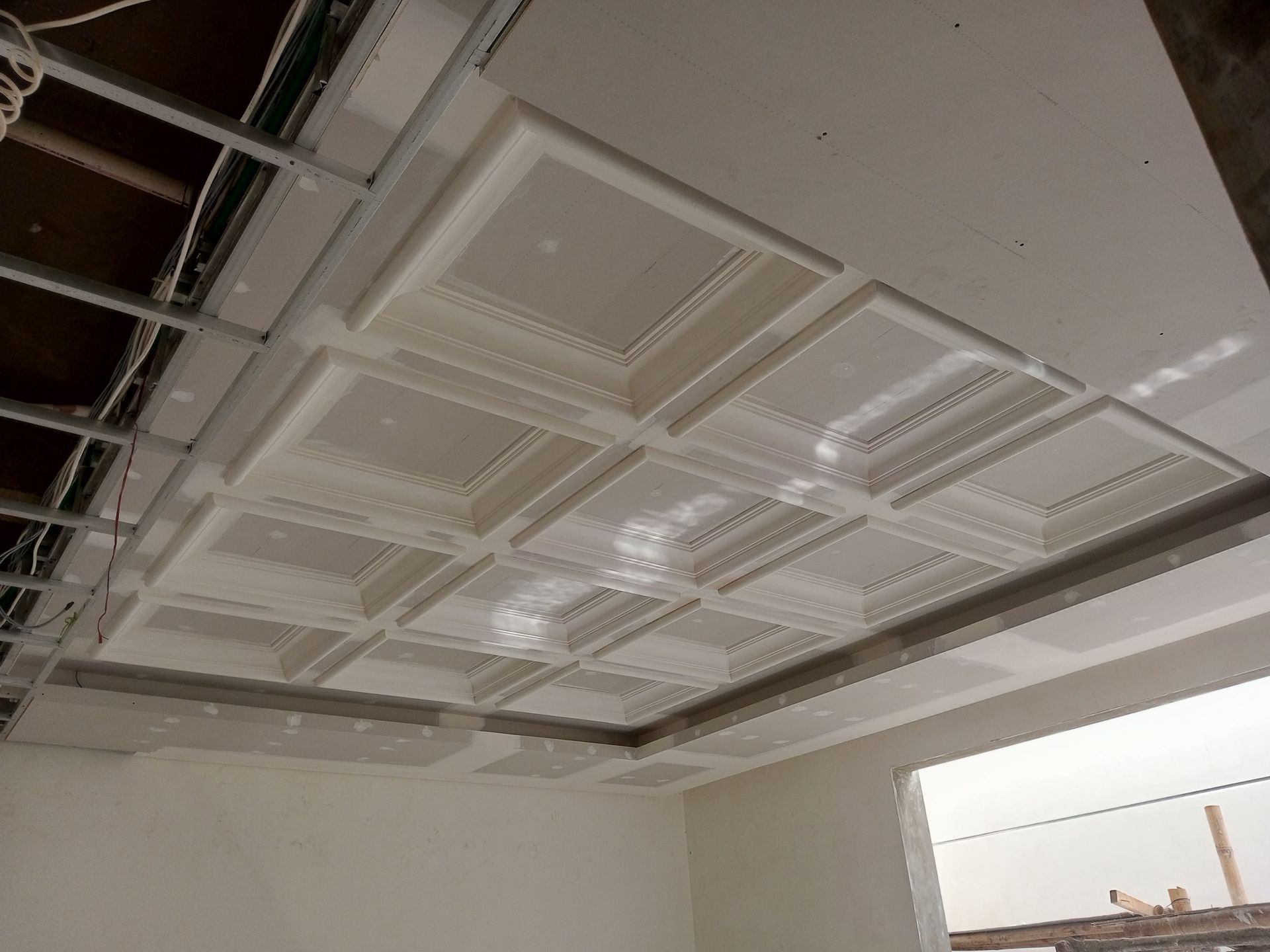 A partially finished ceiling with recessed square panels and metal framing, in a white room with a doorway.