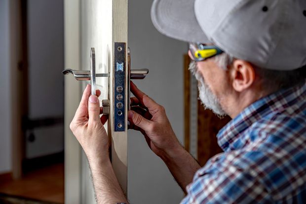 A technician in a plaid shirt and cap installs a metal door handle mechanism into the side of a door.