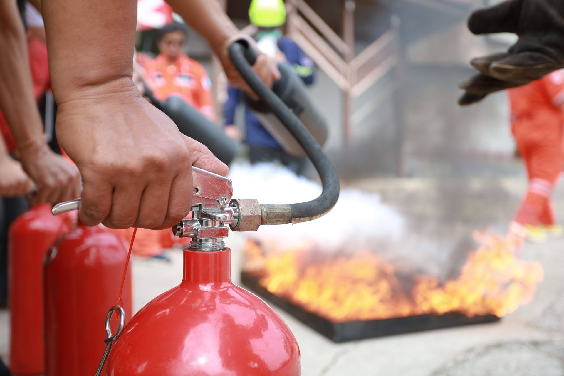 Hands holding a fire extinguisher, spraying foam at a small fire in a square container.