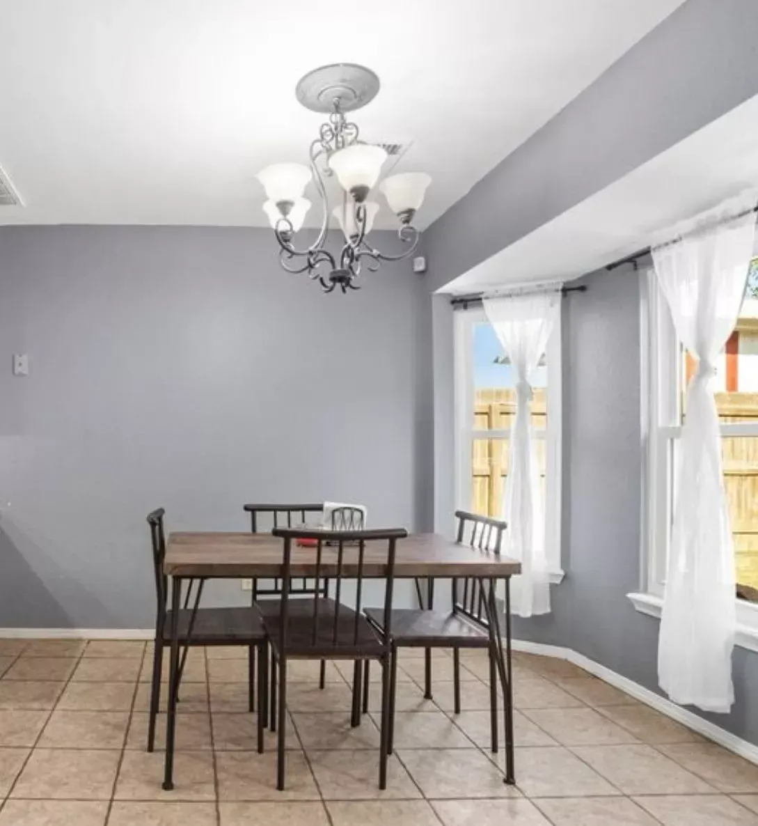 Dining room with table and chairs. Grey walls, chandelier, and bay window with curtains.