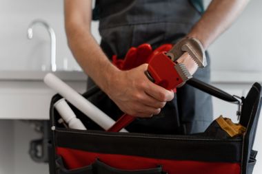 Plumber holding a red pipe wrench, standing in front of a sink, reaching into a tool bag.