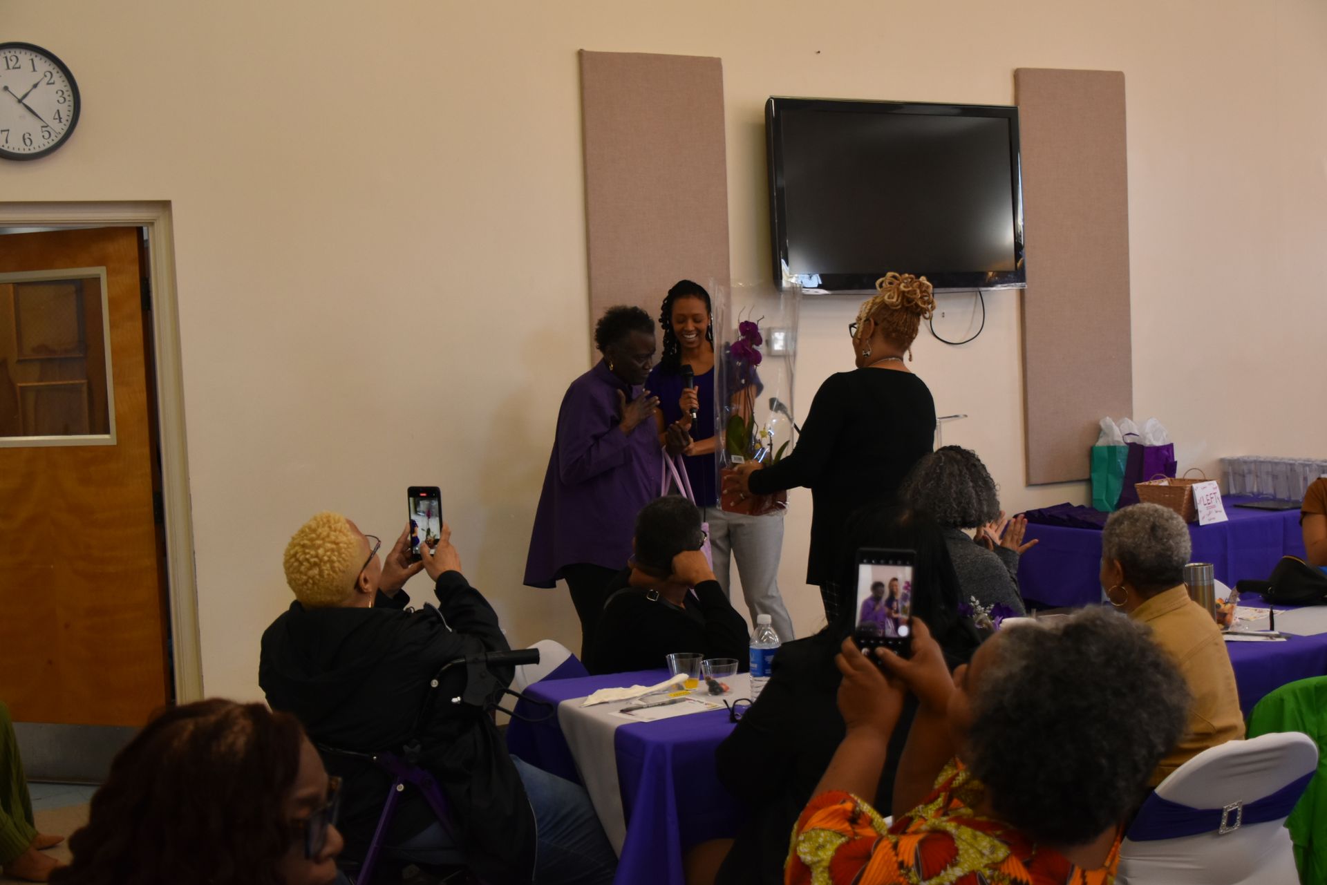 A group of people are sitting at tables in a room with a clock on the wall.