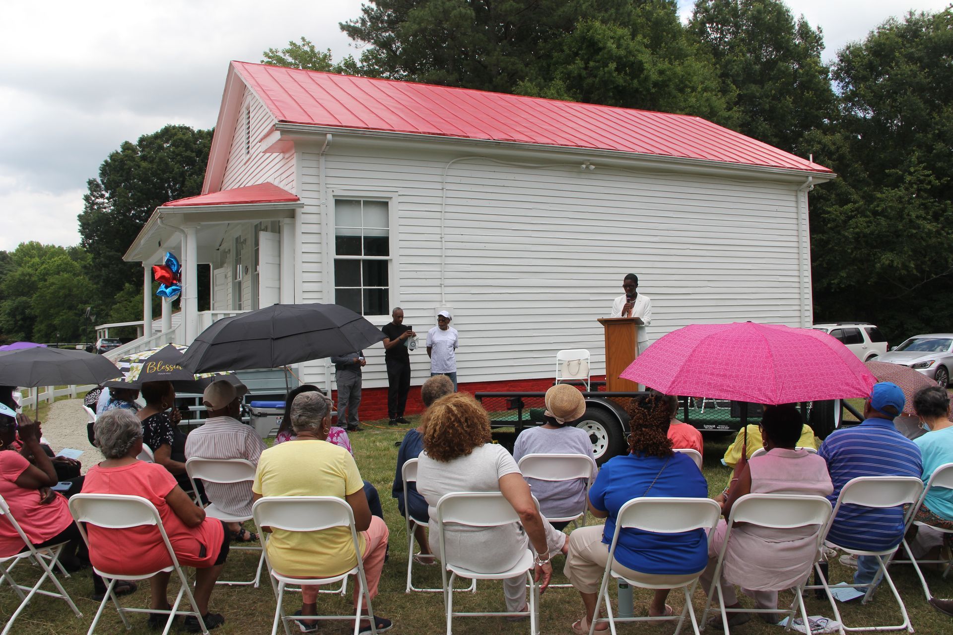 A group of people are sitting in chairs in front of a white building with a red roof.