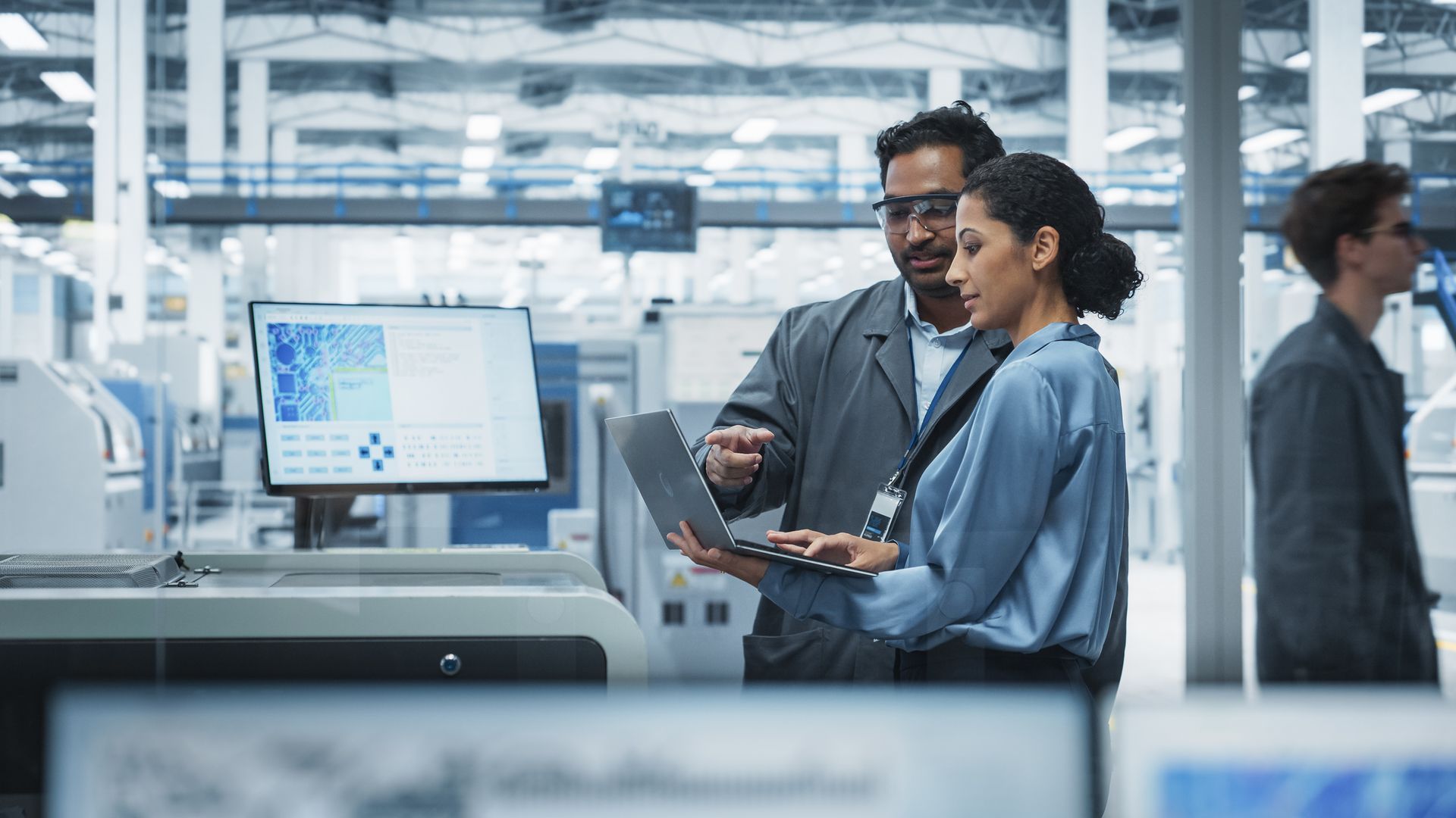 Two people reviewing data on a laptop and screen in a factory setting.