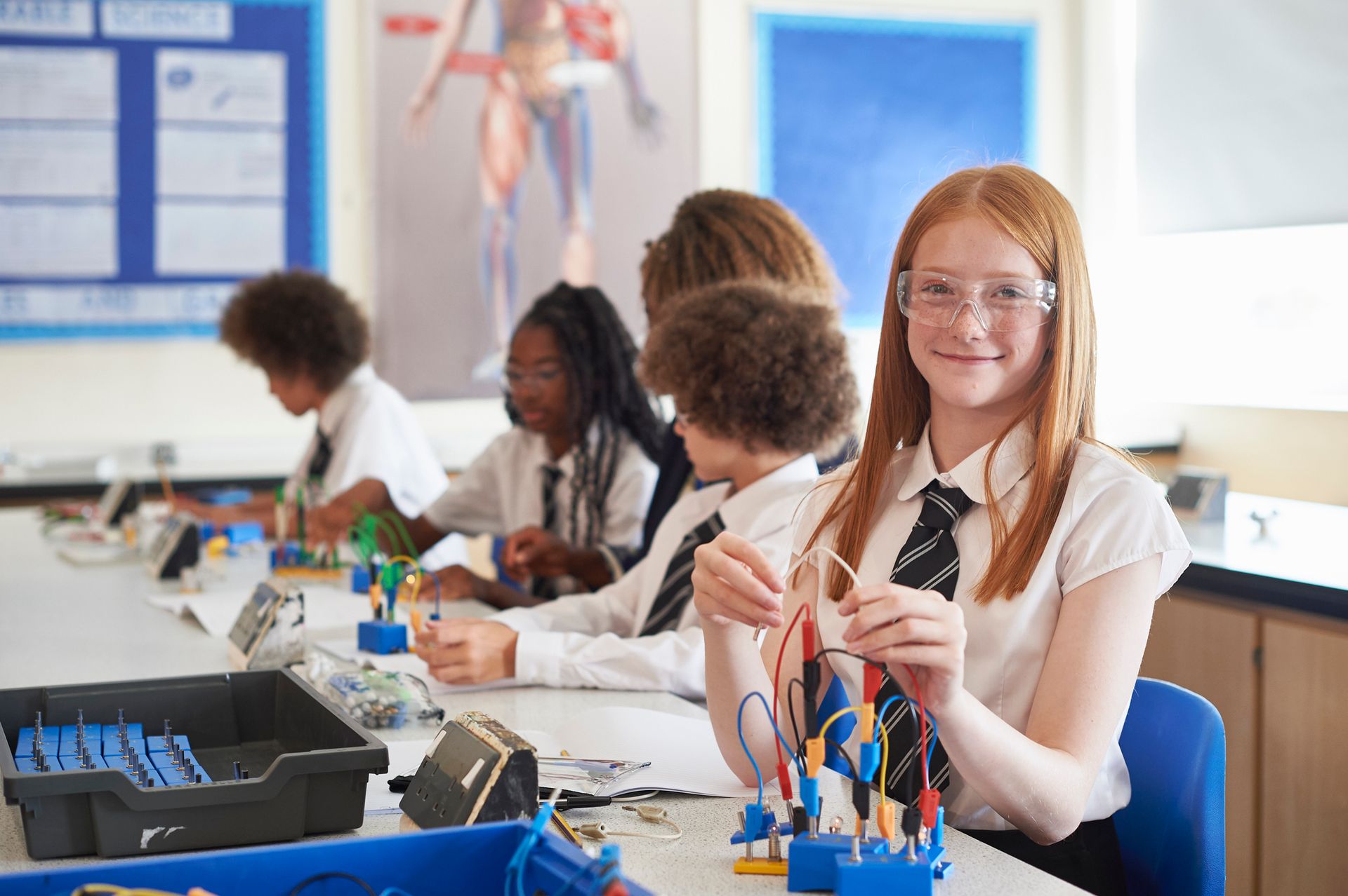 Students in school uniforms in a science lab; one smiles at the camera while working with wires.