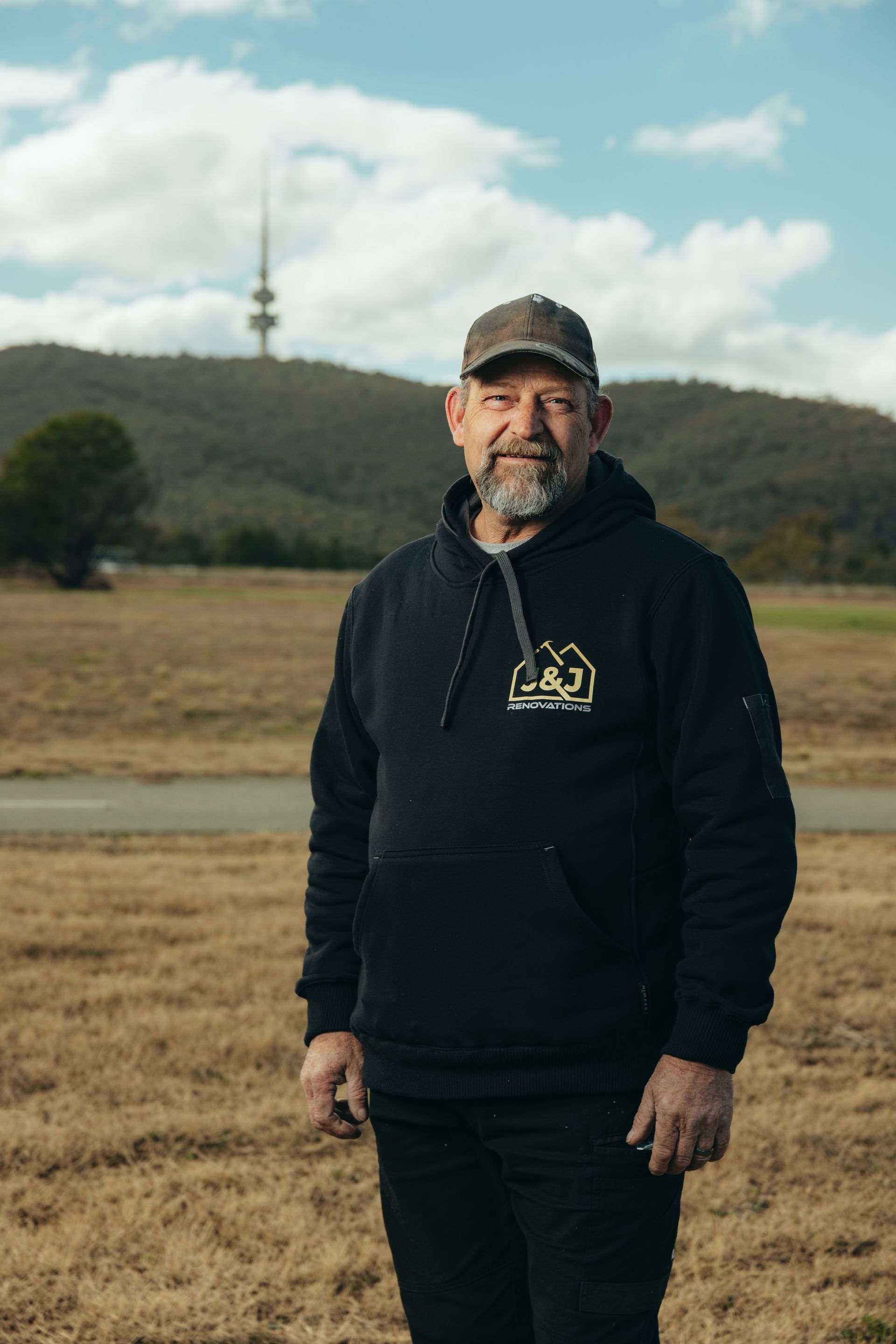 Man in cap and hoodie stands in field with distant tower and hill.