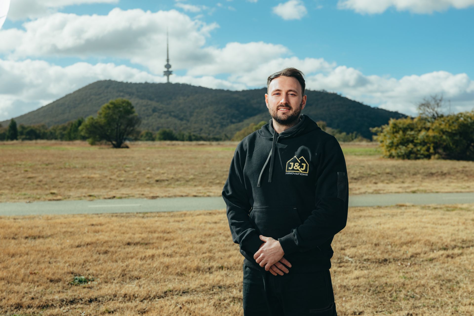 Man in black hoodie standing in front of a grassy field, a mountain, and a tower on a sunny day.
