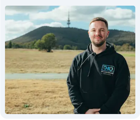 Man in black hoodie, smiling, stands outdoors with a hill and tower in the background.