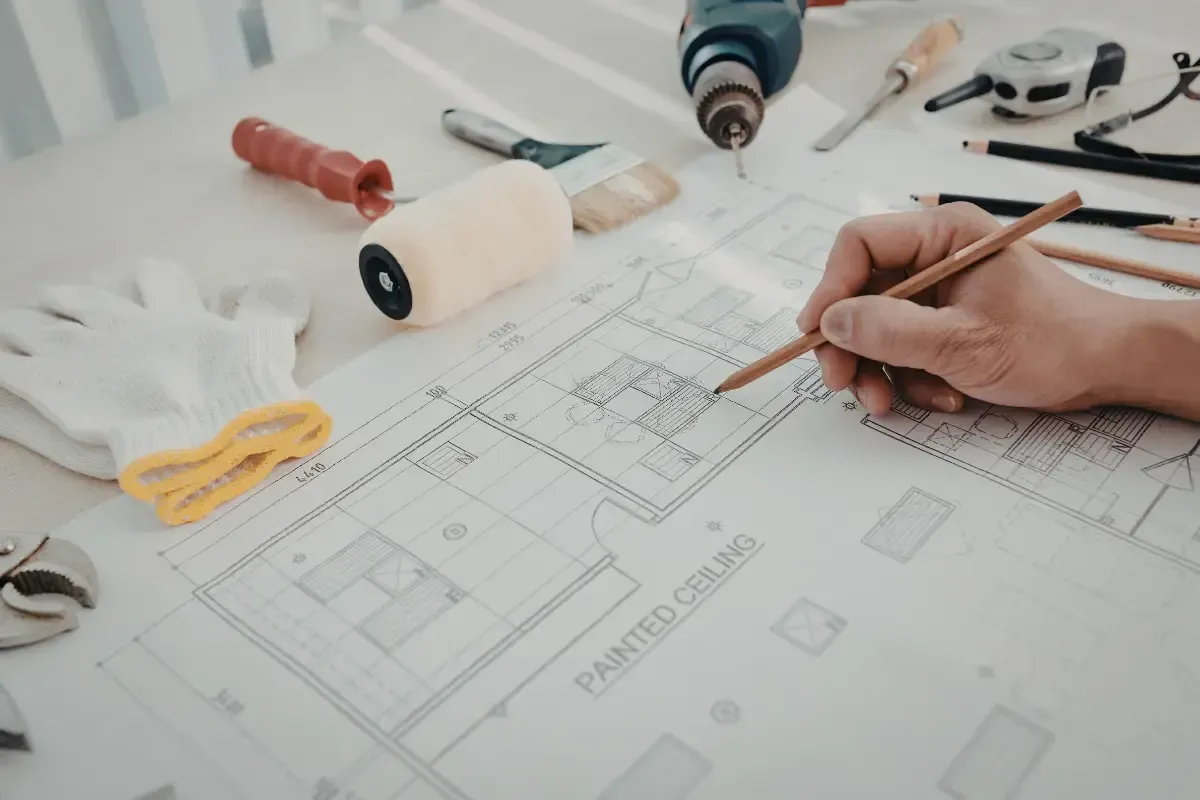 Person's hand sketching on a blueprint with tools, including a drill, roller, and gloves, on a white surface.