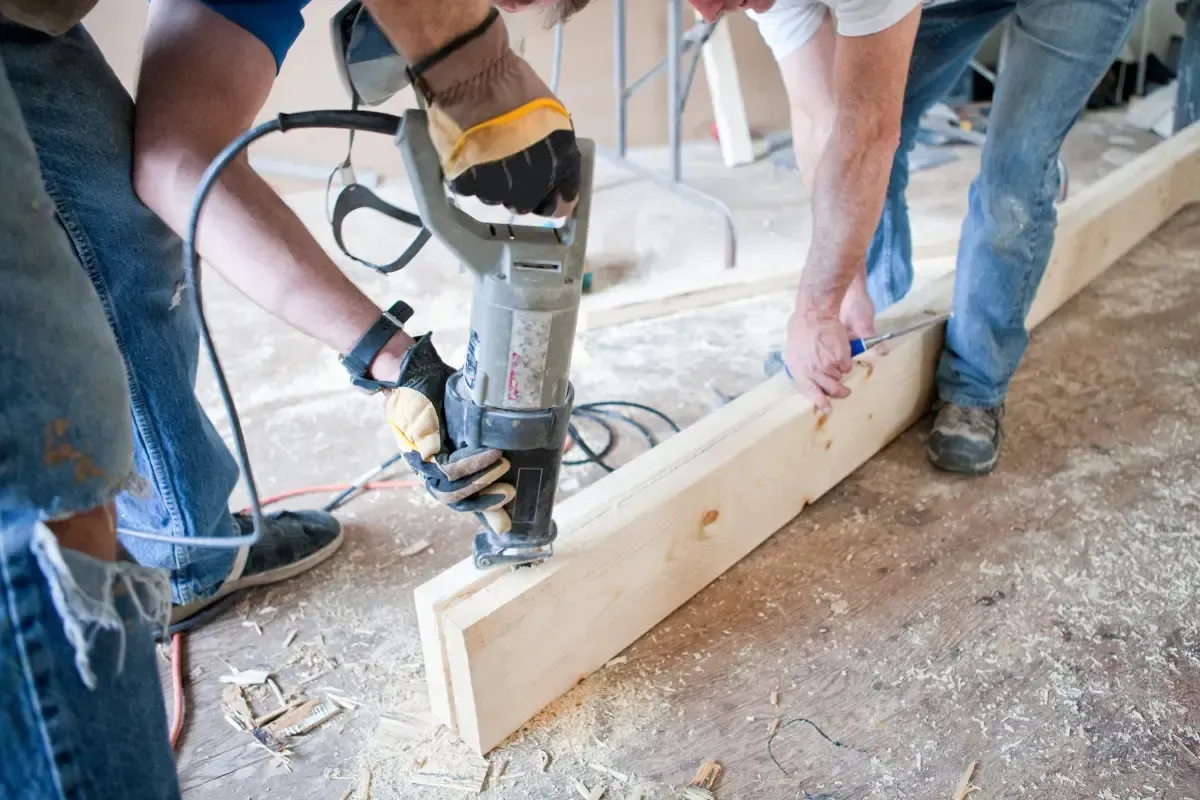 People using a power tool to work on a wooden beam on a floor.