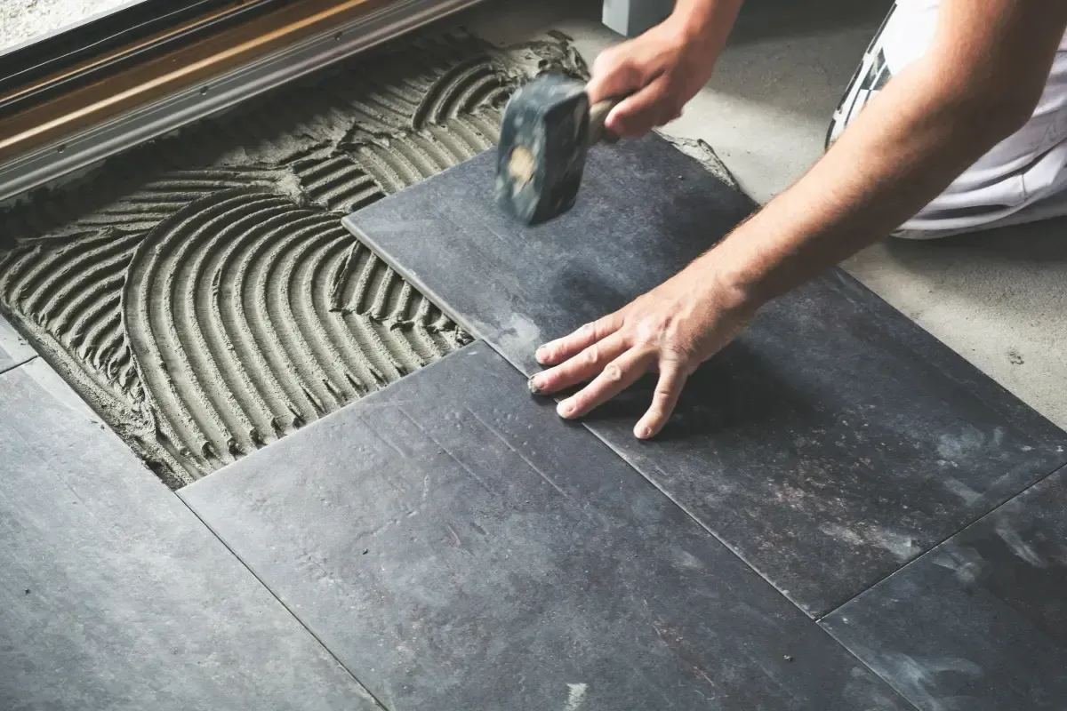 A person uses a rubber mallet to level a dark grey floor tile onto a freshly troweled adhesive bed.
