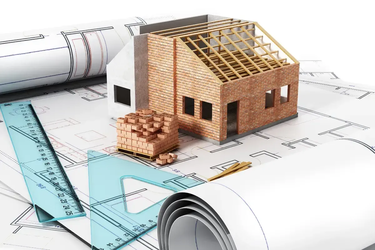 Model house under construction on architectural blueprints with ruler and rolled paper.