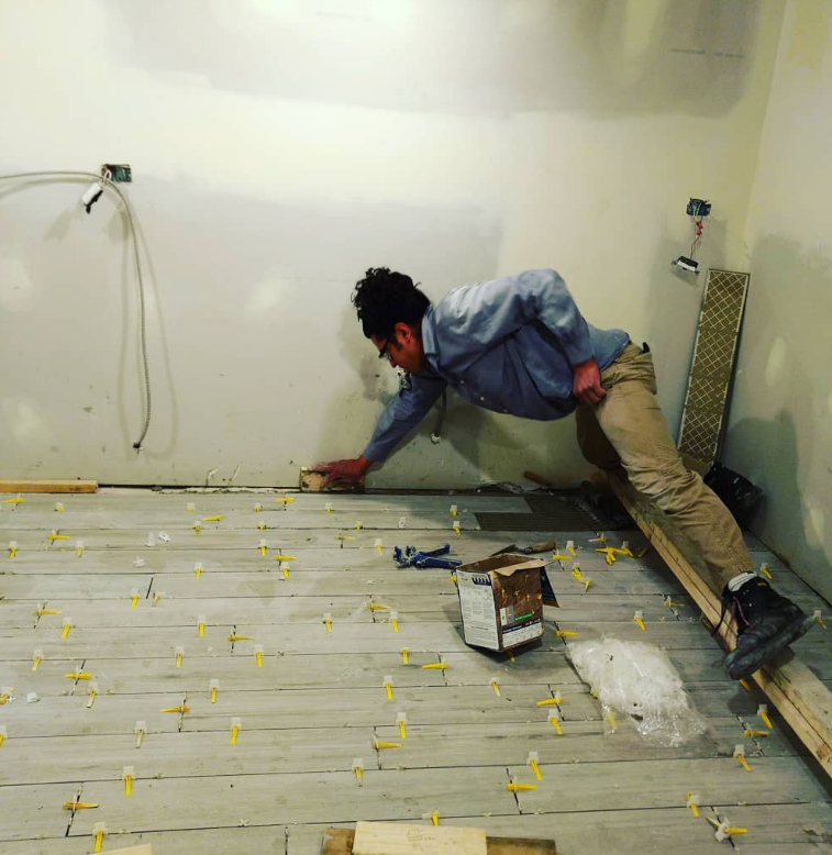 Person tiling a floor, on hands and knees, in a room under construction, with tiles and leveling spacers.