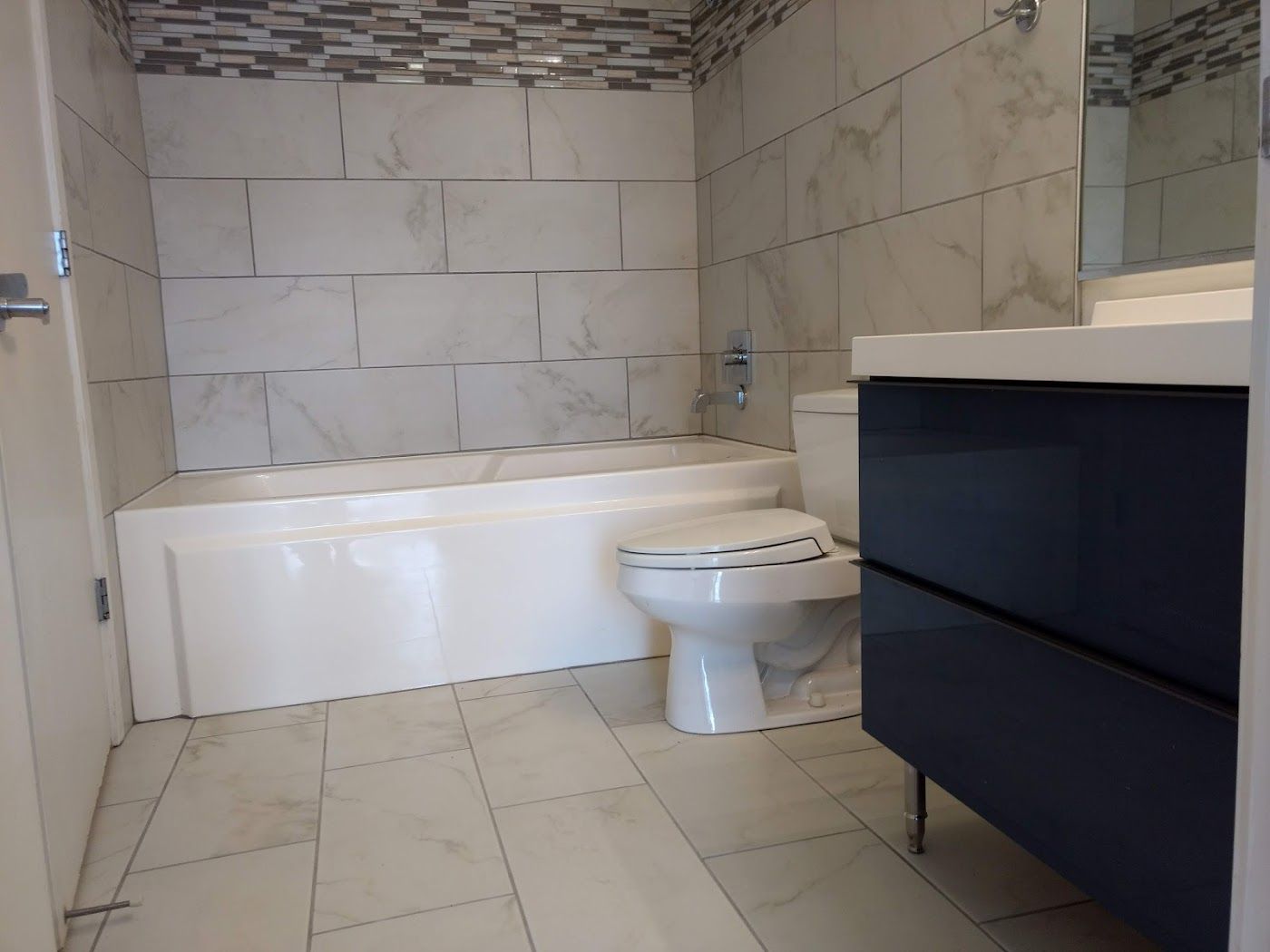 Modern bathroom with a white tub, toilet, navy vanity, and marble-like tile.