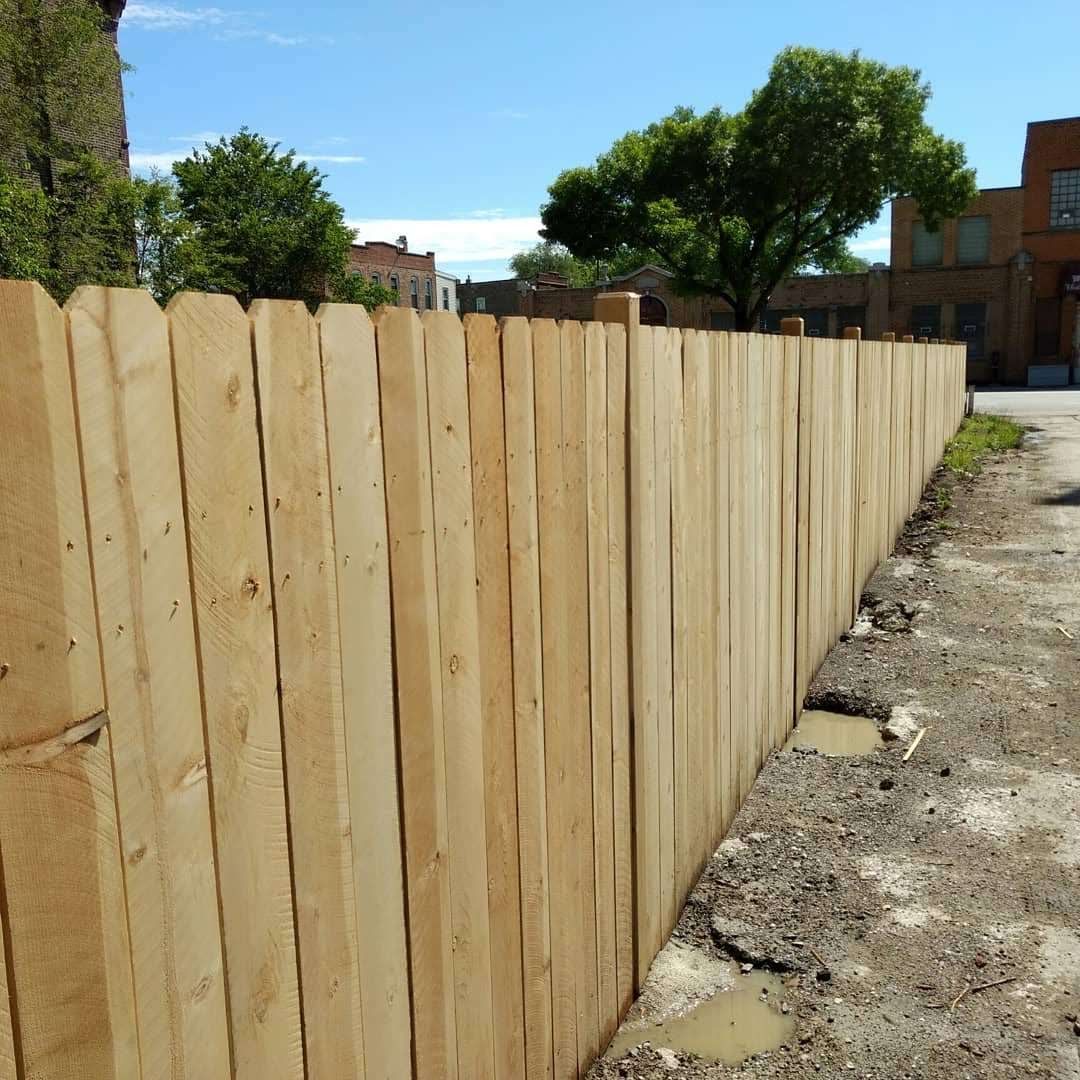 Wooden fence along a gravel path with buildings and trees in the background under a blue sky.