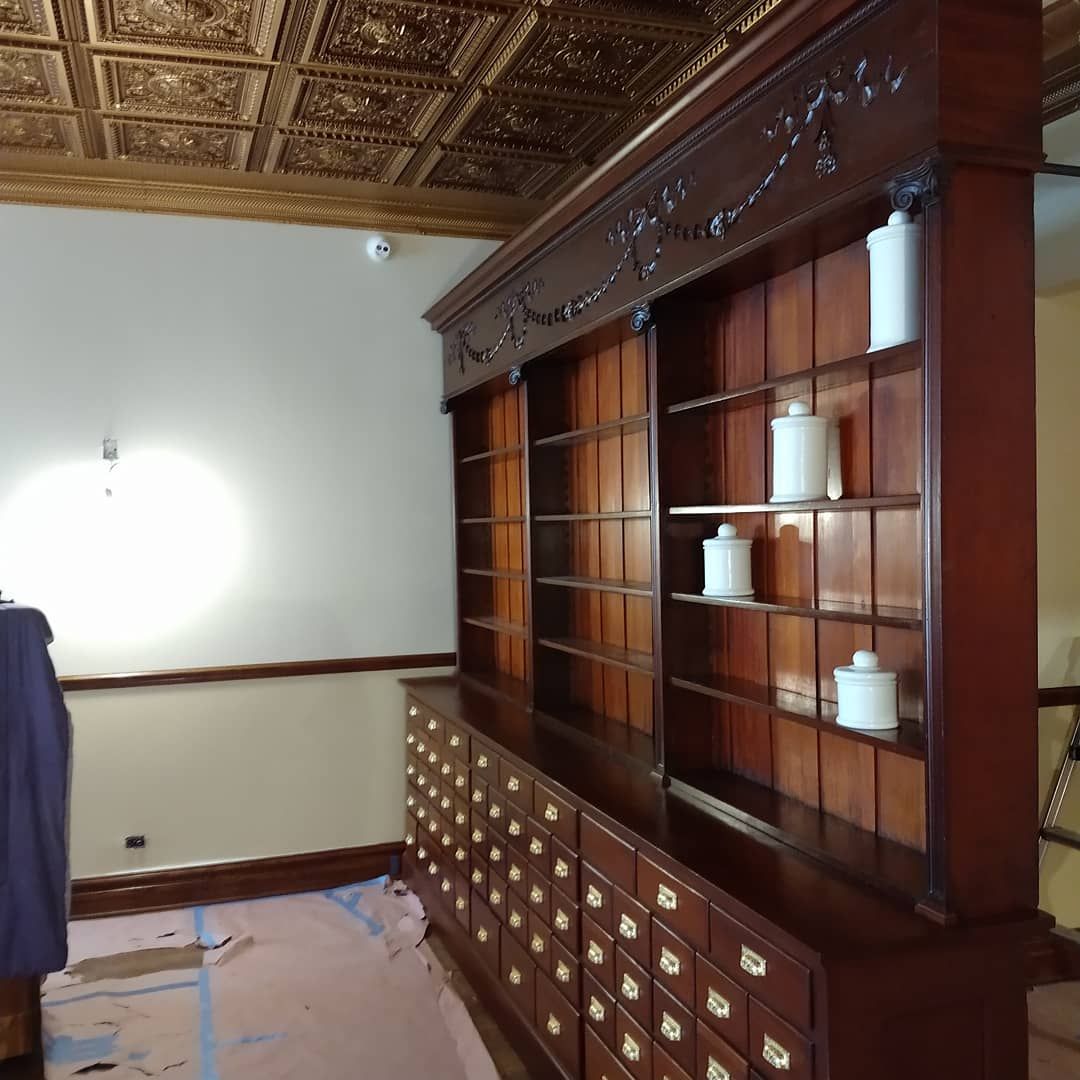 Wooden cabinet with drawers and shelves; ornate ceiling and walls; white jars on shelves.
