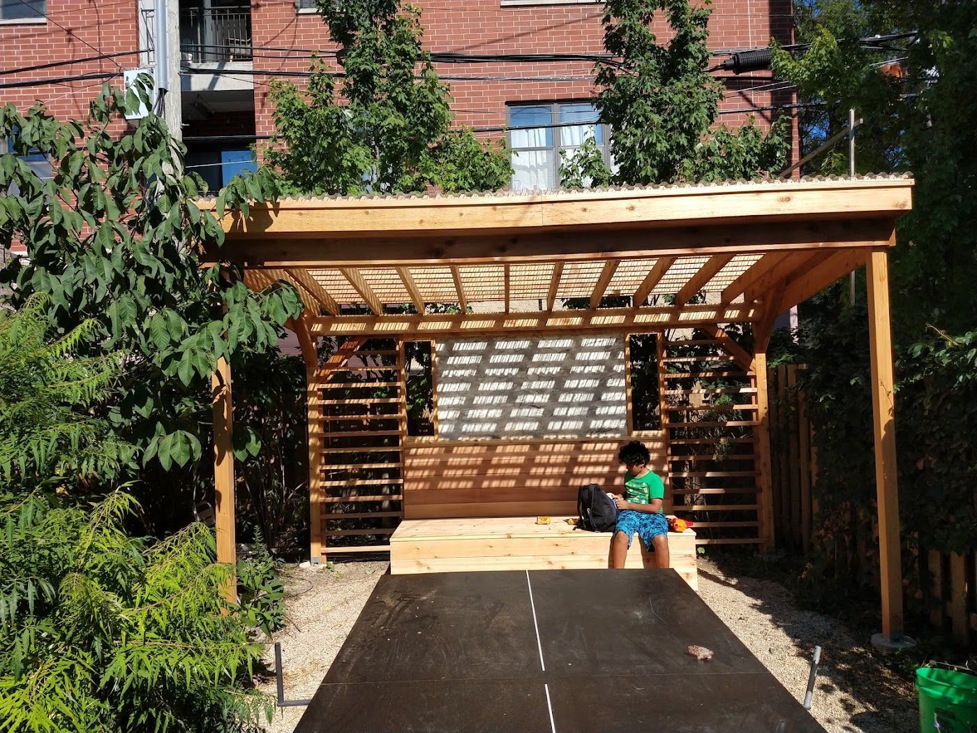 Wooden pergola with slatted walls and roof, partially covered with greenery, person inside.