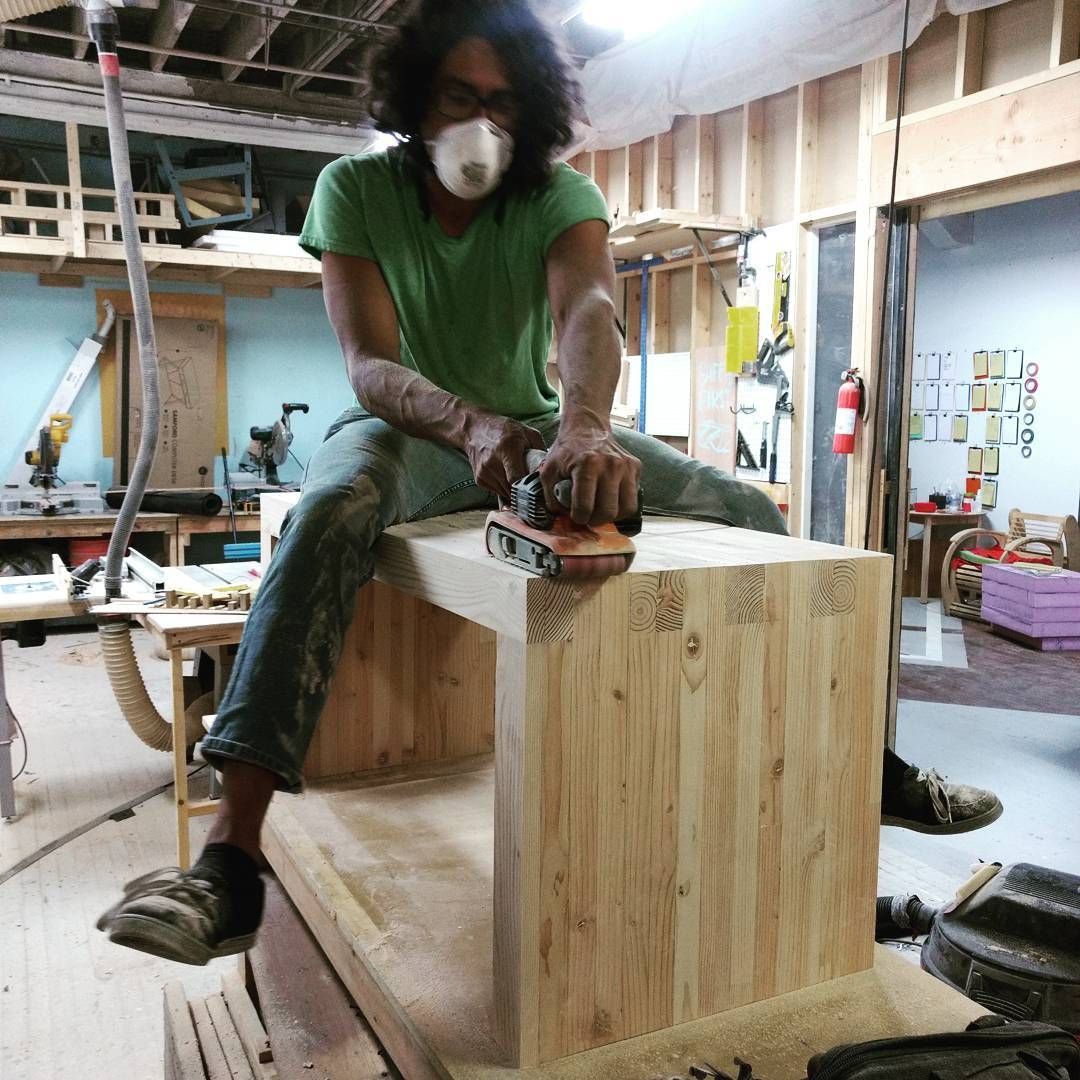 Person sanding a wooden bench in a workshop, wearing safety mask, green shirt, and jeans, sitting on the bench.