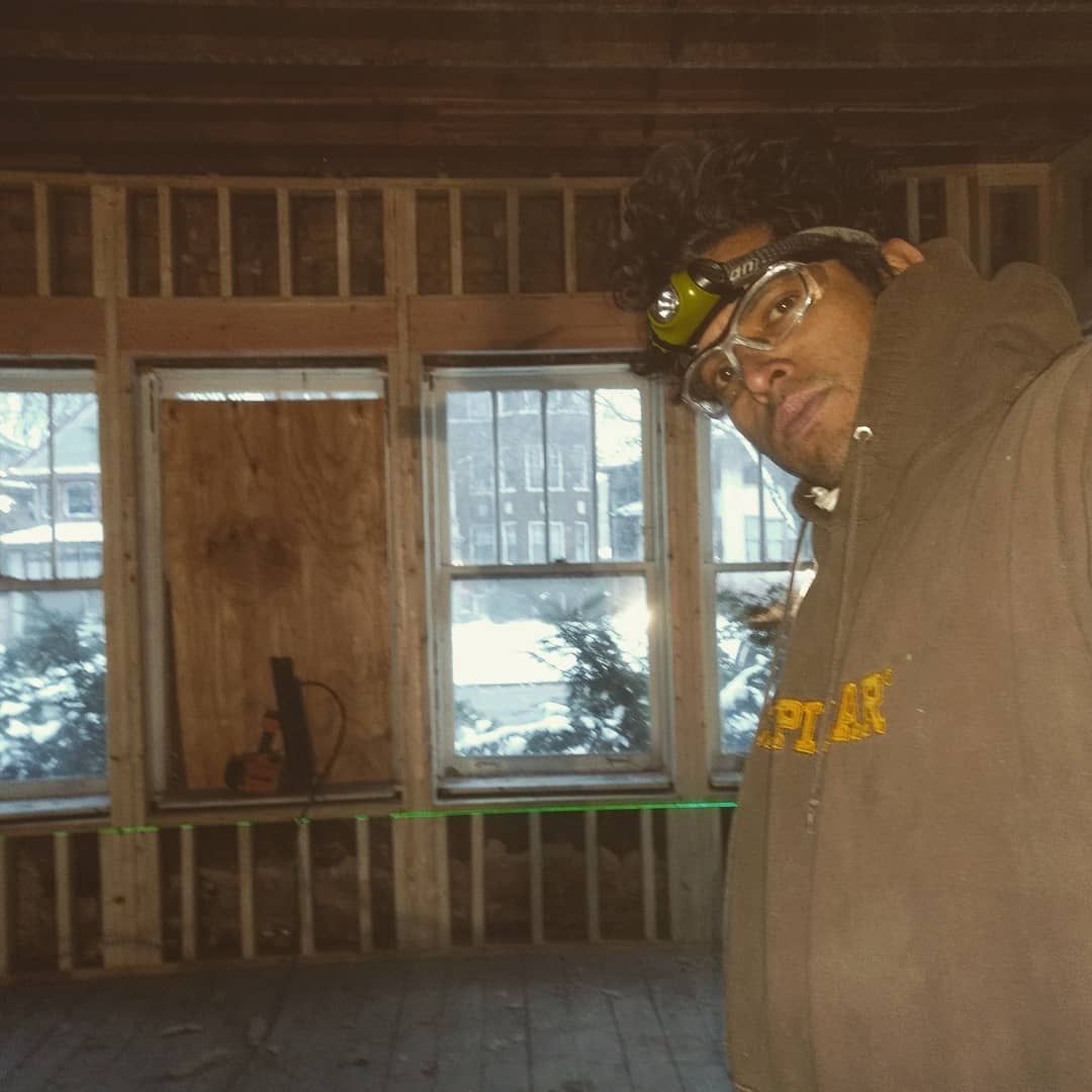 Man in brown hoodie and safety glasses in a room with exposed framing, looking at the camera.