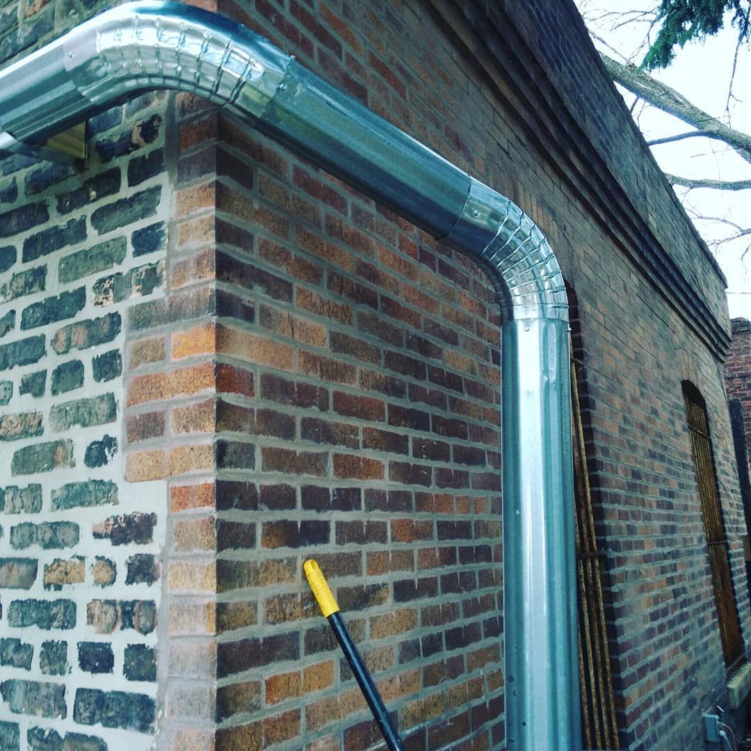 Silver metal rain gutter on brick building's corner. Broom with yellow handle leans against the wall.
