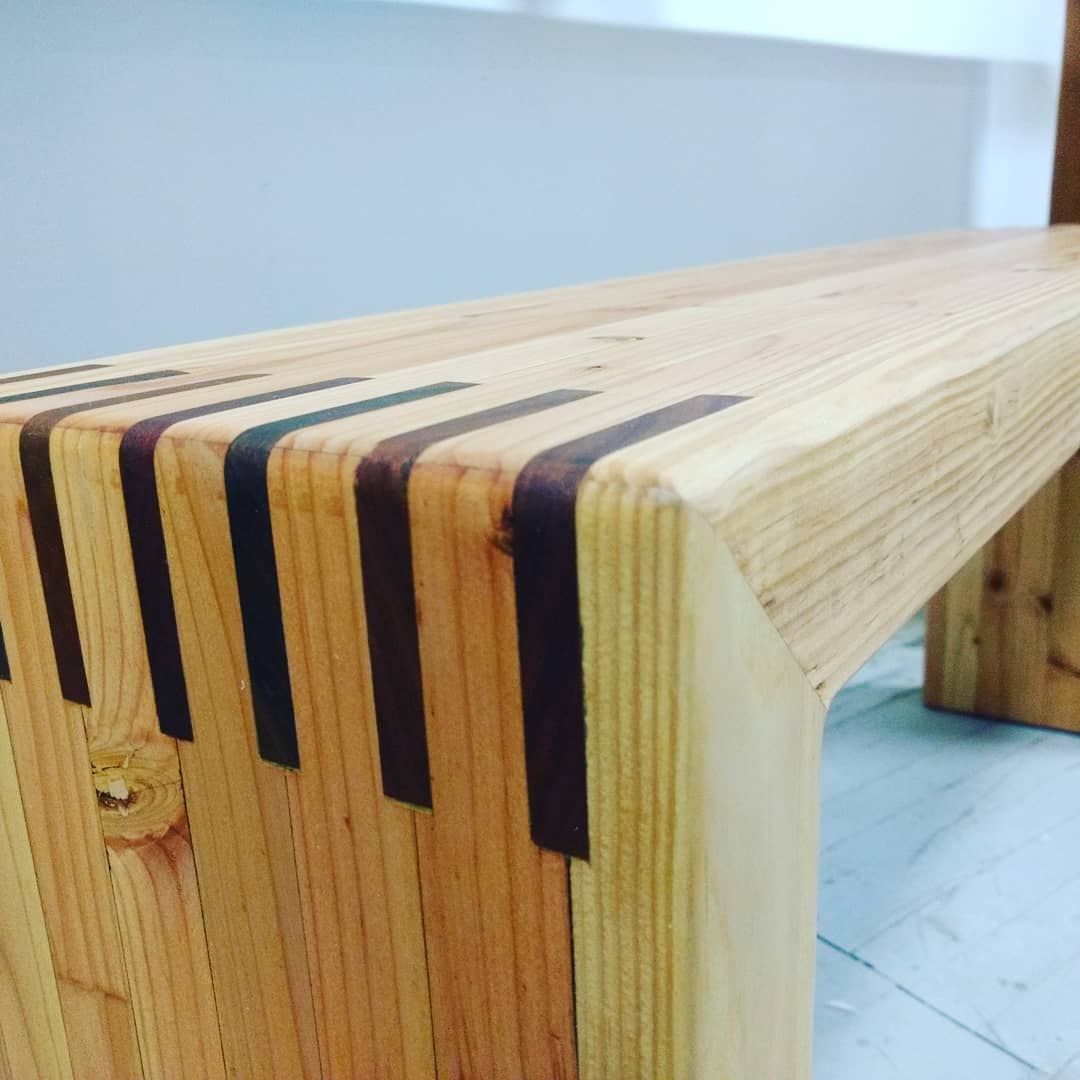 Wooden bench with dark brown inlays along the top and corner. Light wood with a white wall backdrop.