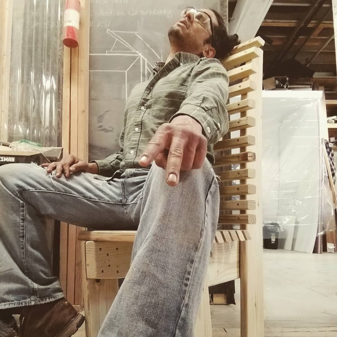 Man sitting in a wooden chair, leaning back, pointing finger. Indoors with metal and wood structure in the background.