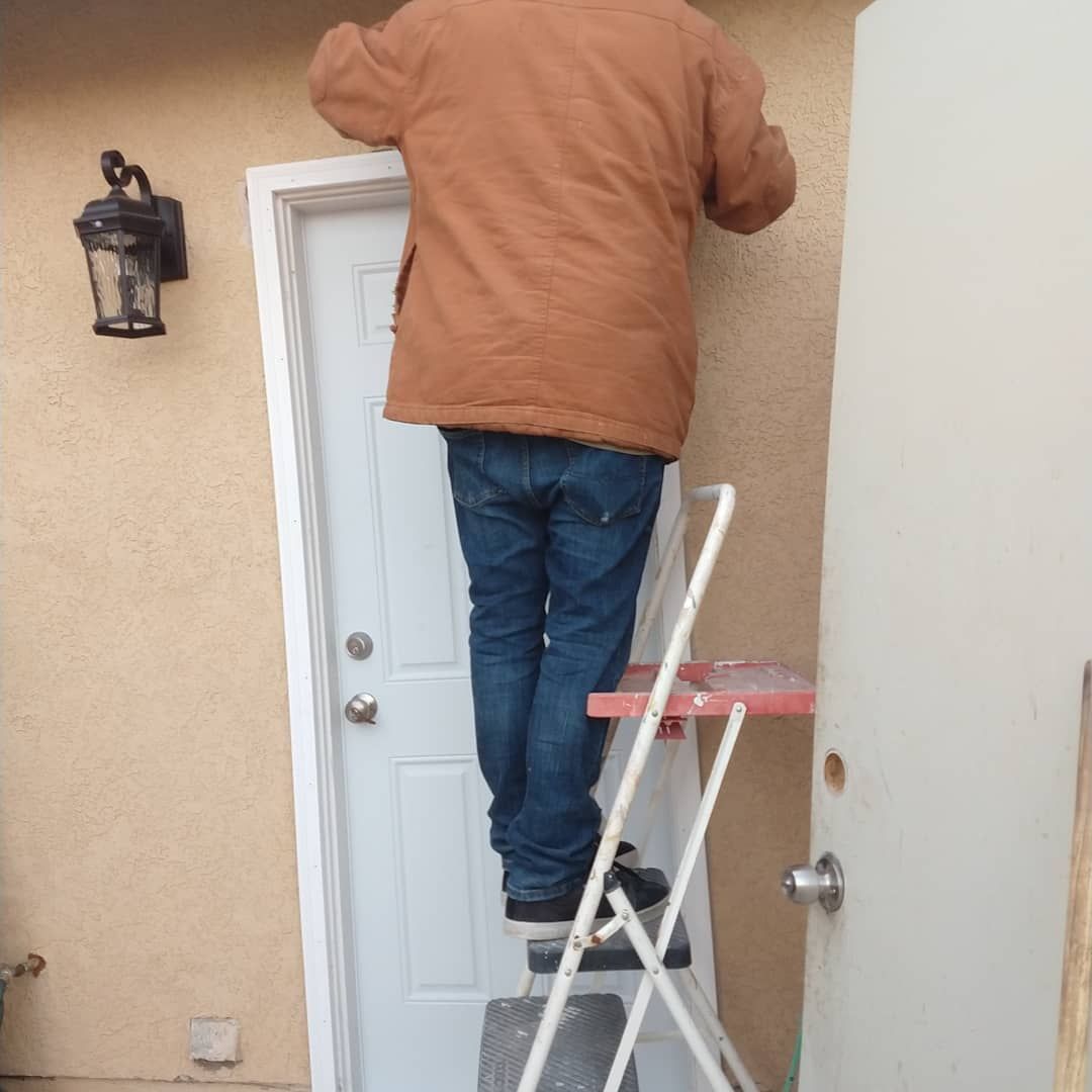 Person on a stepladder working above a doorway outdoors, wearing a brown jacket and jeans.