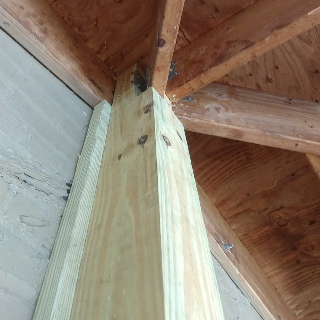 Wooden post supporting a roof's interior framing. Light wood beams and a vertical post are visible against a white wall.