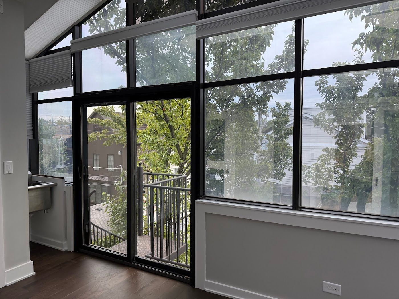 Interior room with large windows, dark frame sliding door to balcony, trees visible outside.