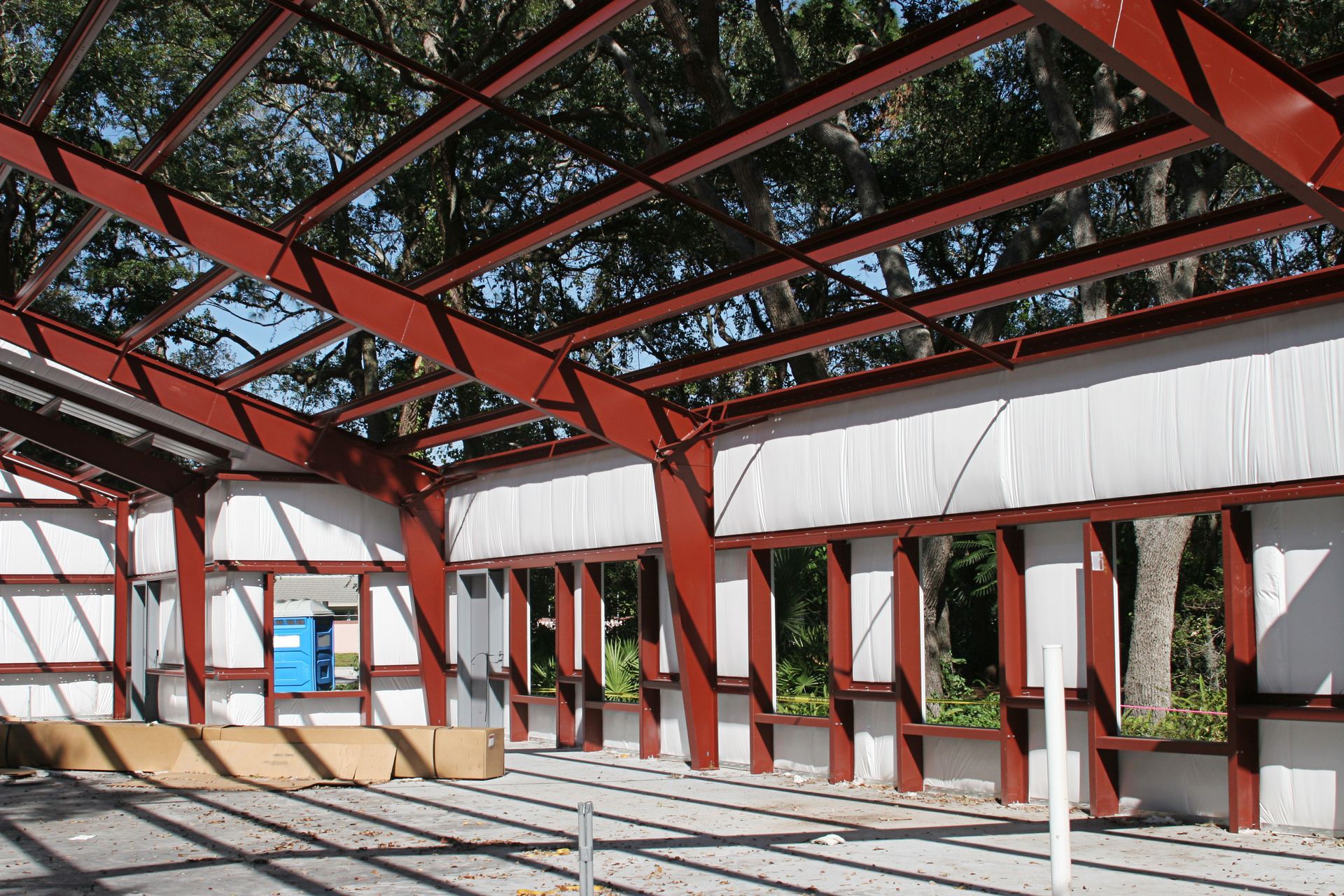 Steel frame of a building under construction, red beams and columns, white walls, sunny outdoors.