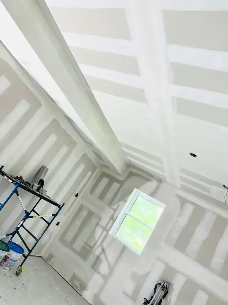 Interior room under construction, drywall installed on walls and ceiling, with a window and scaffolding.