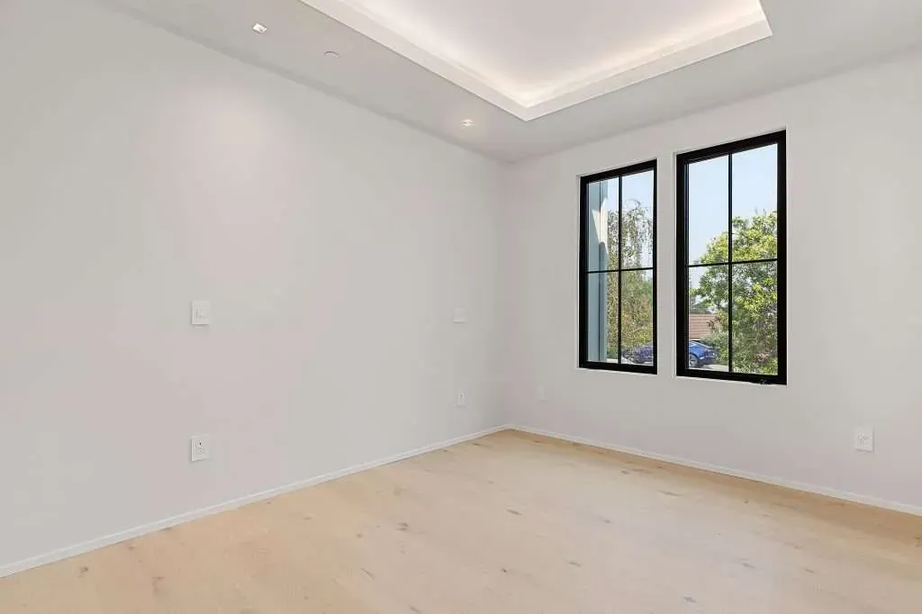 Empty room with light wood floors, white walls, and two black-framed windows overlooking greenery.