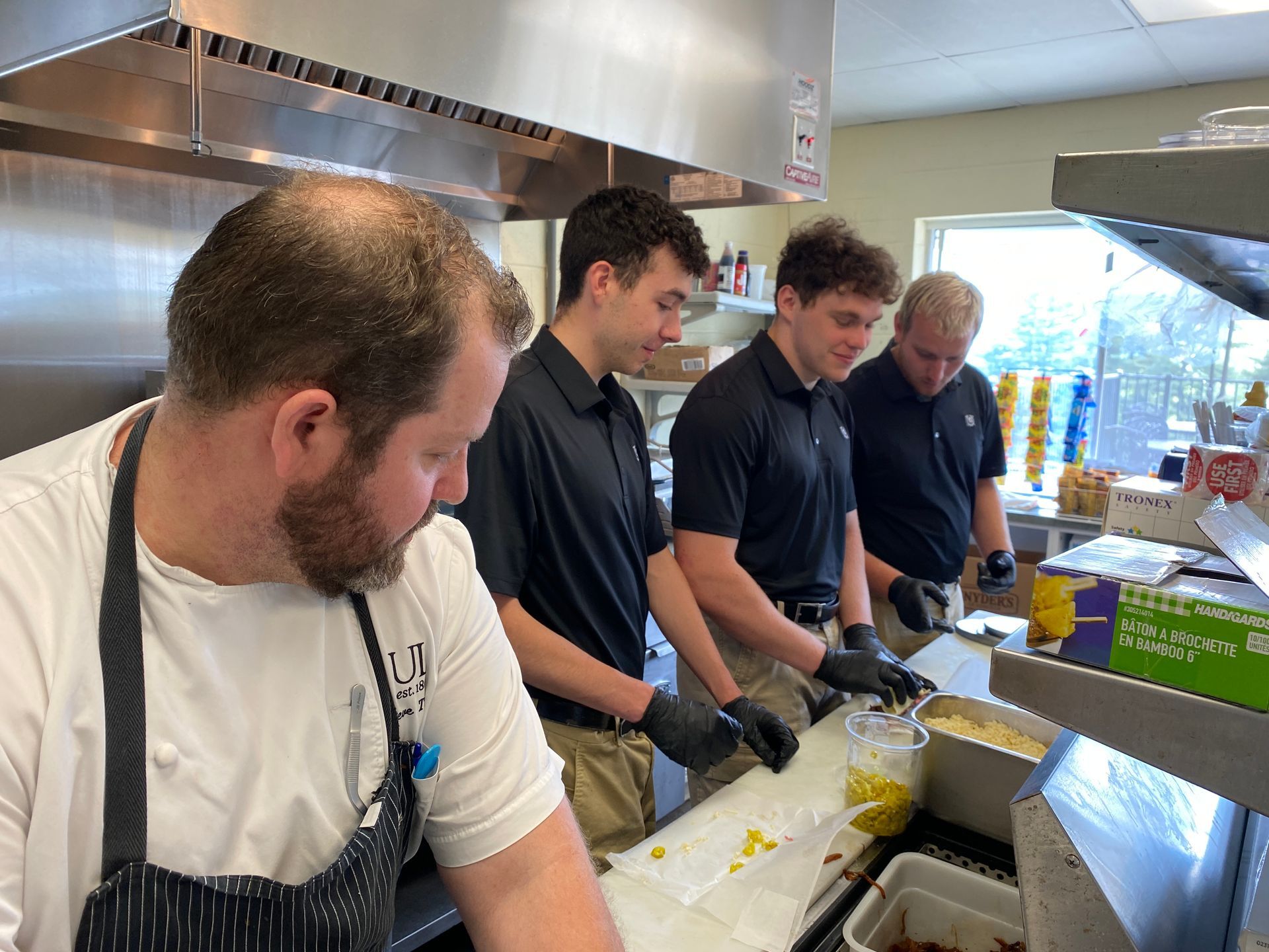 Chef and three young men prepare food in a kitchen. They wear aprons and gloves, focused on their task.