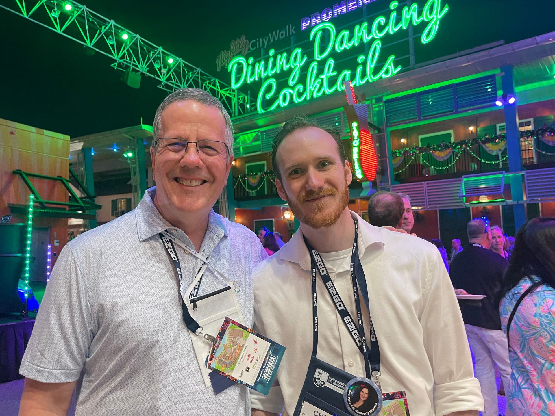 Two men smiling in front of a neon sign that reads 