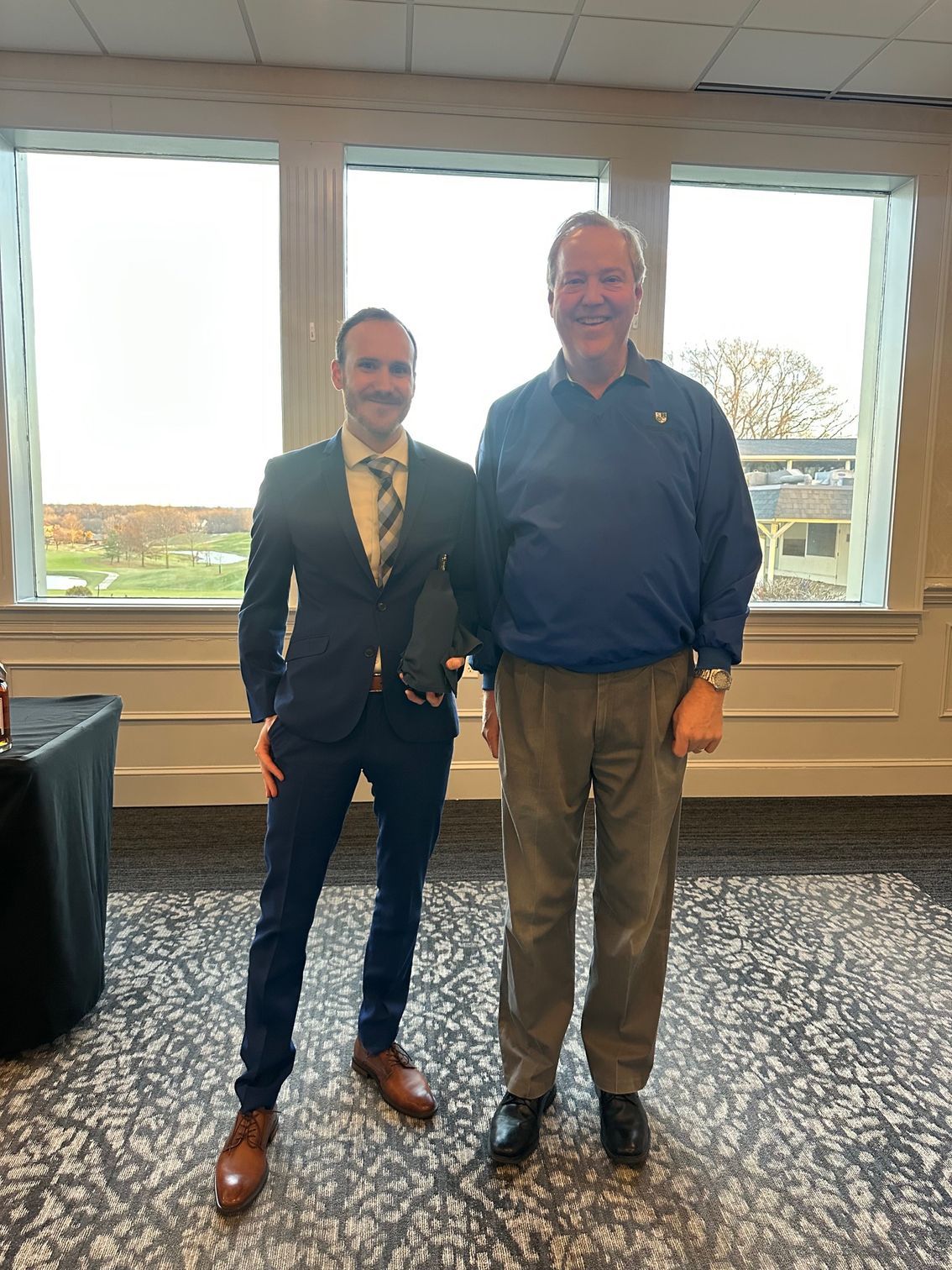 Two men pose near windows, one in a suit holding a trophy, the other in a sweater and khakis.