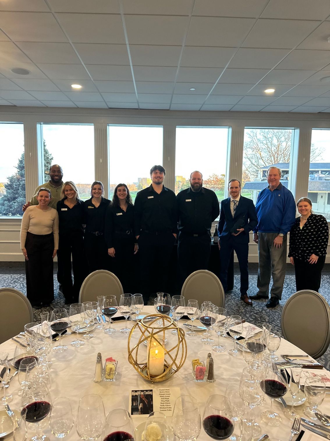 Group of people standing behind a decorated table, with a window backdrop.