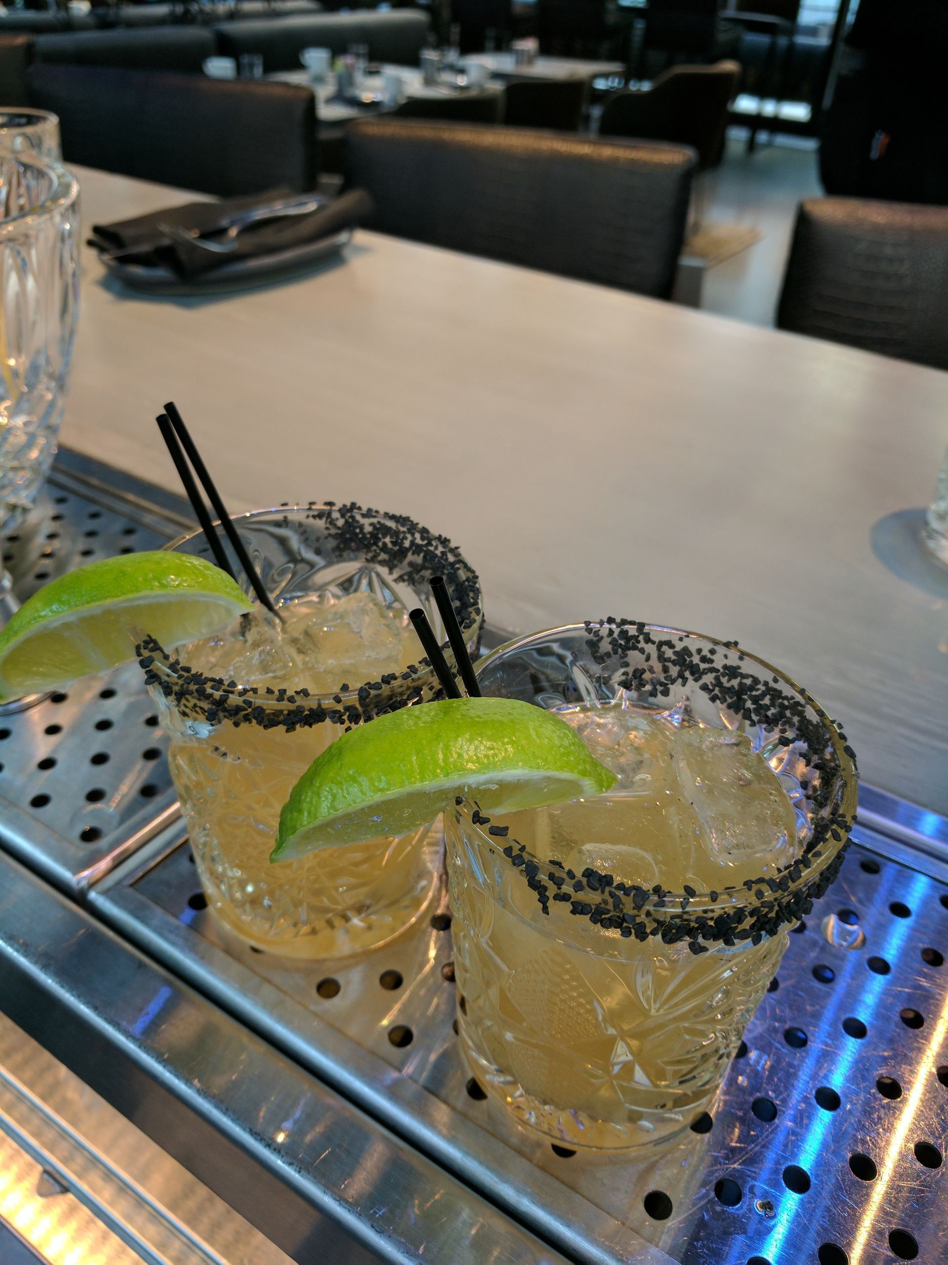 Two margaritas with black salt rims, lime wedges, and straws on a bar.