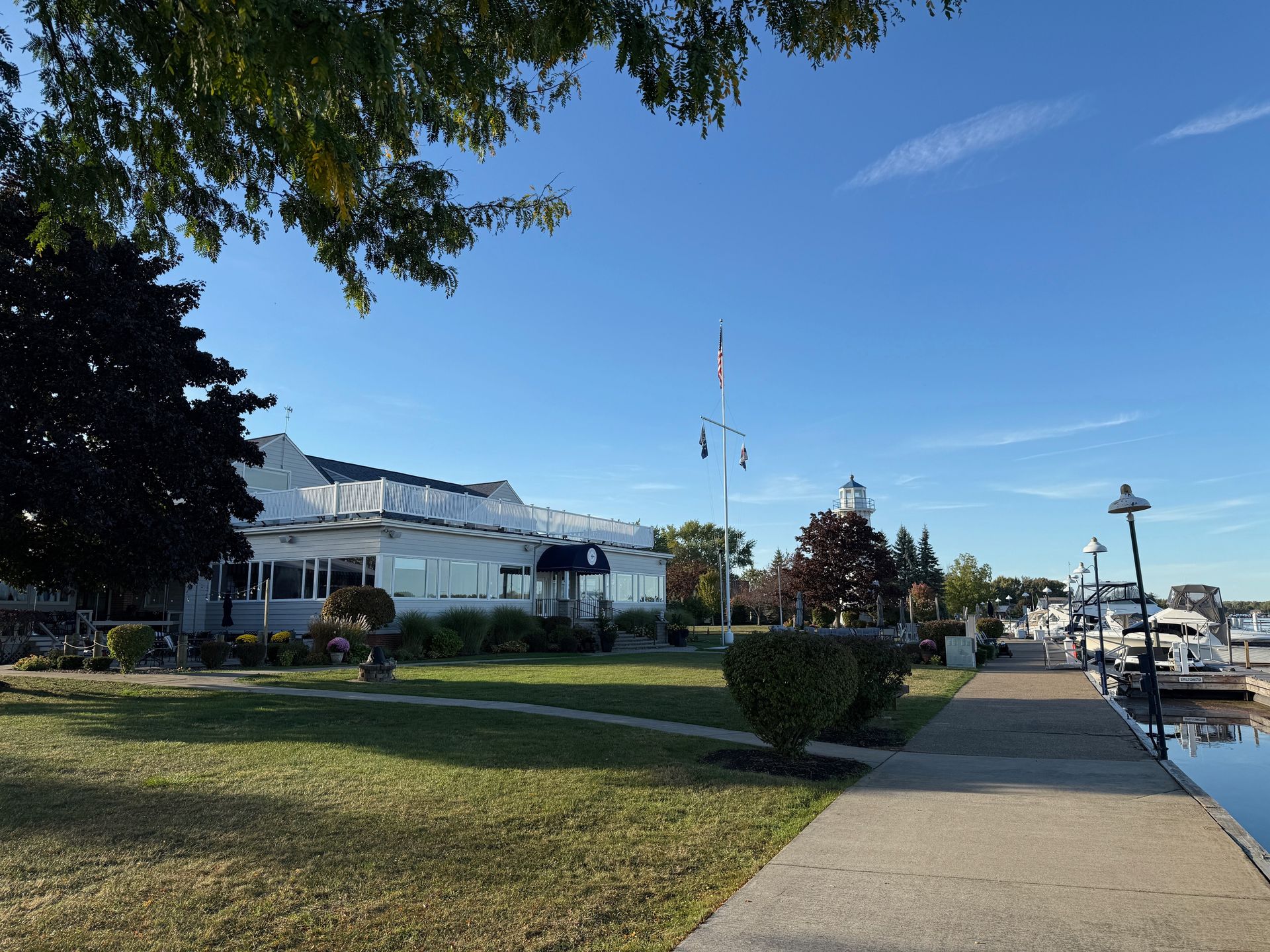 Lakeside building with a flag and docked boats. Paved path and grassy area under a blue sky.