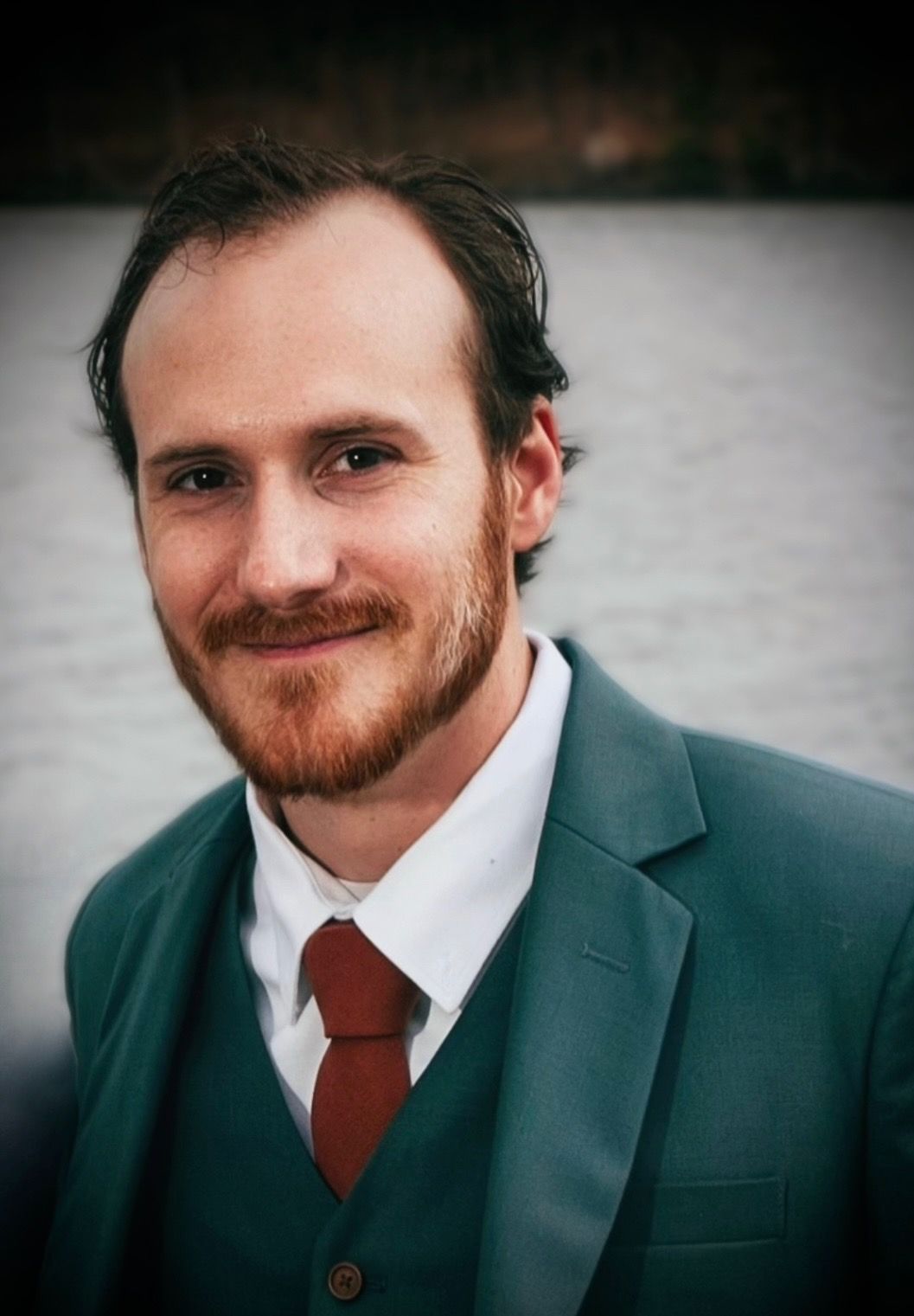 Man in teal suit and red tie smiles, facing camera with body of water in background.