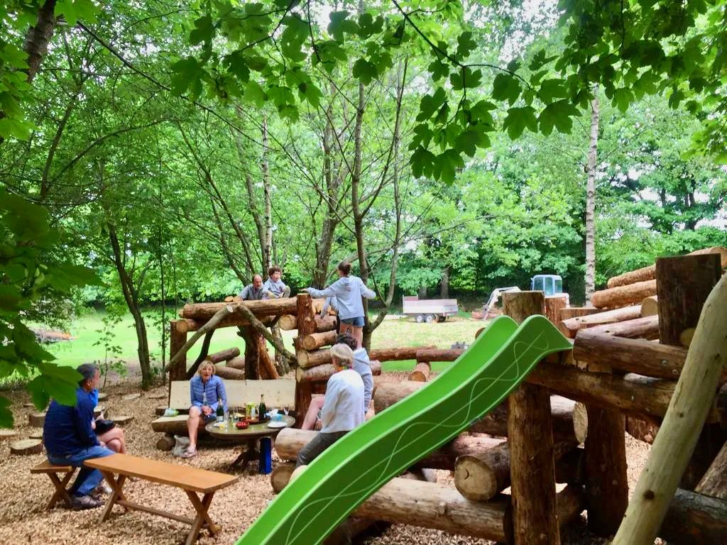 Een groep mensen zit aan een picknicktafel in het bos, vlak bij een glijbaan.